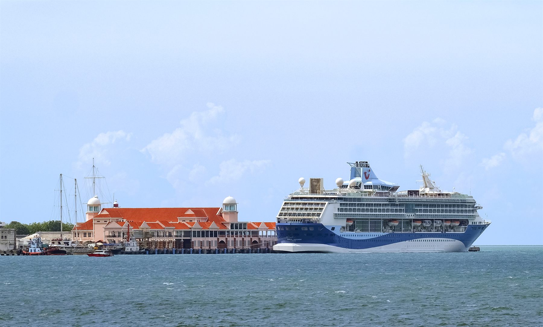 A cruise ship docked beside a colonial-style pier building with red roofs